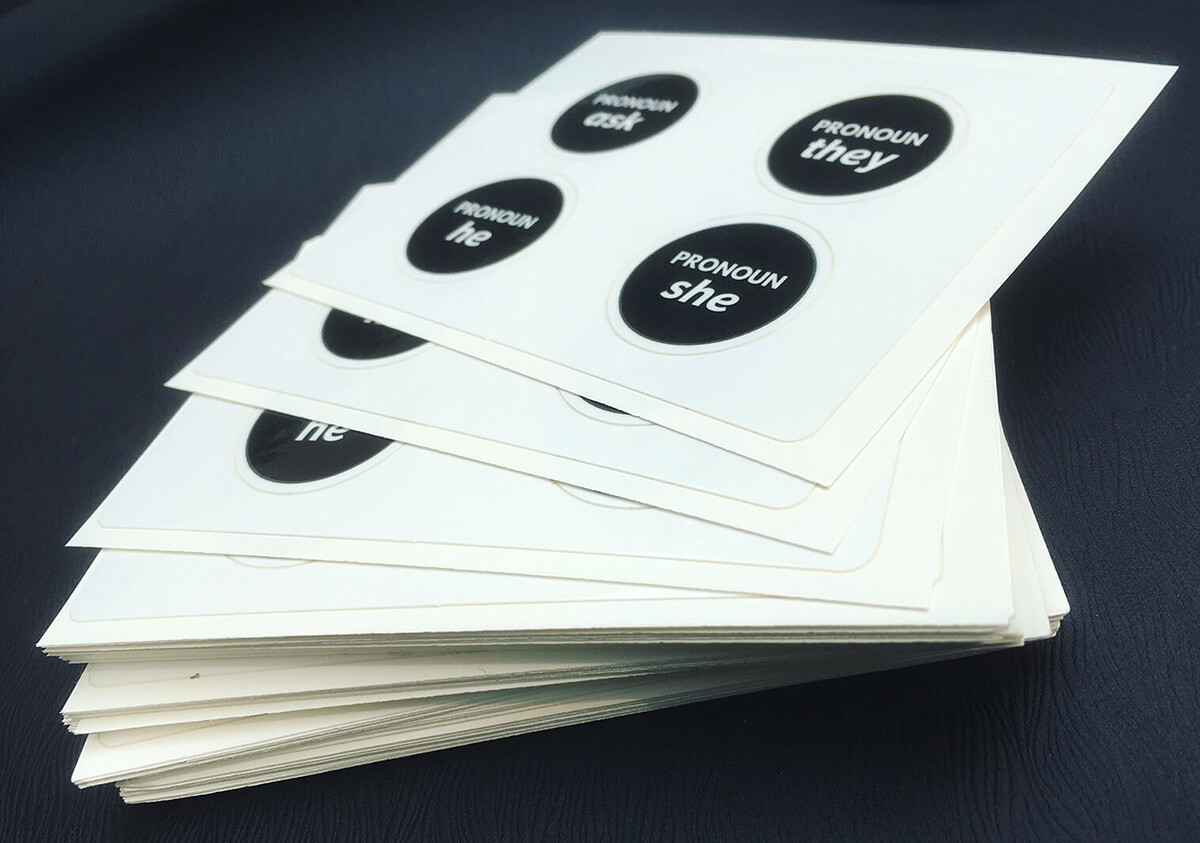 Fanned out stack of sticker sheets; each sheet features black stickers with ask, they, he, and she as pronoun options