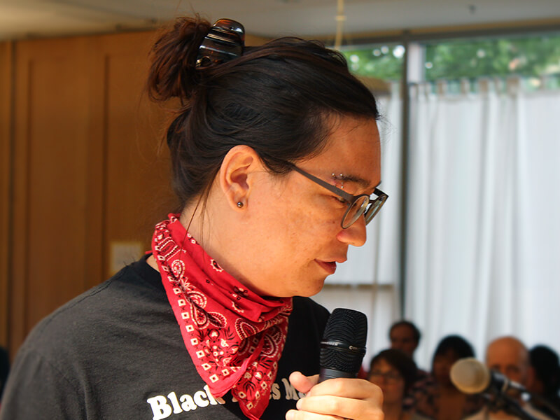 Closeup of Emily Gorcenski turned to the right and holding a mic while pausing mid-talk. They are wearing a Black Lives Matter shirt and in front of an audience.