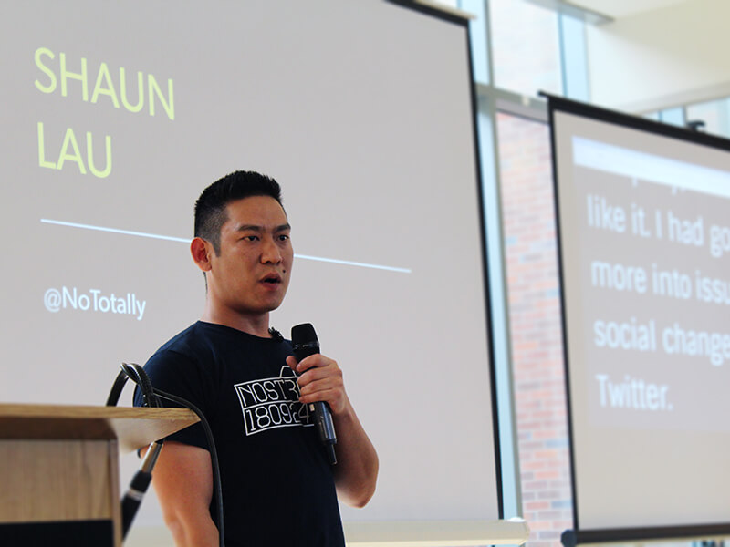 Photo of Shaun Lau standing and talking seriously into a mic. Behind him is a slide displaying his name.